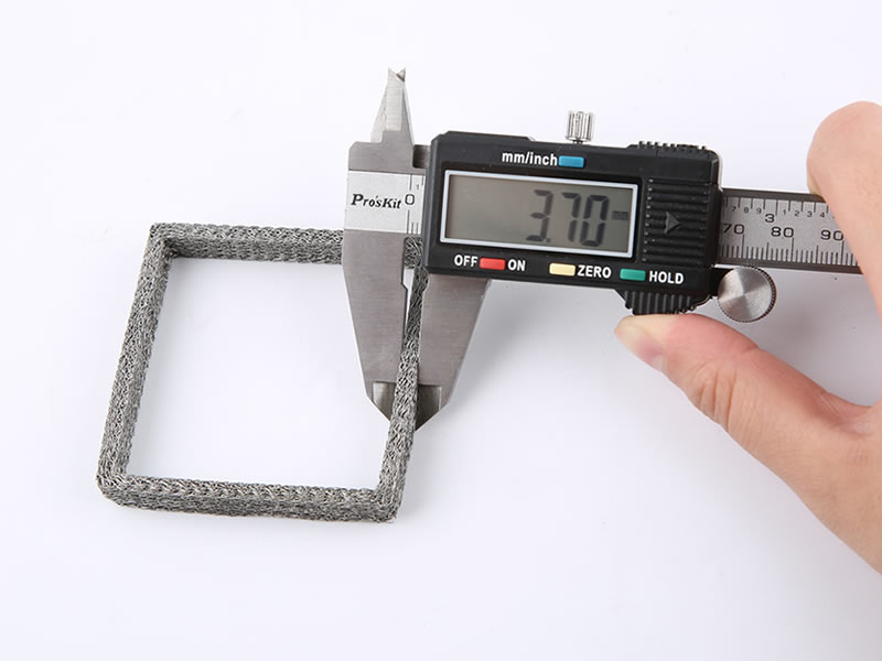 Una mano mide el grosor de la malla de punto comprimida rectangular mediante un calibrador vernier.