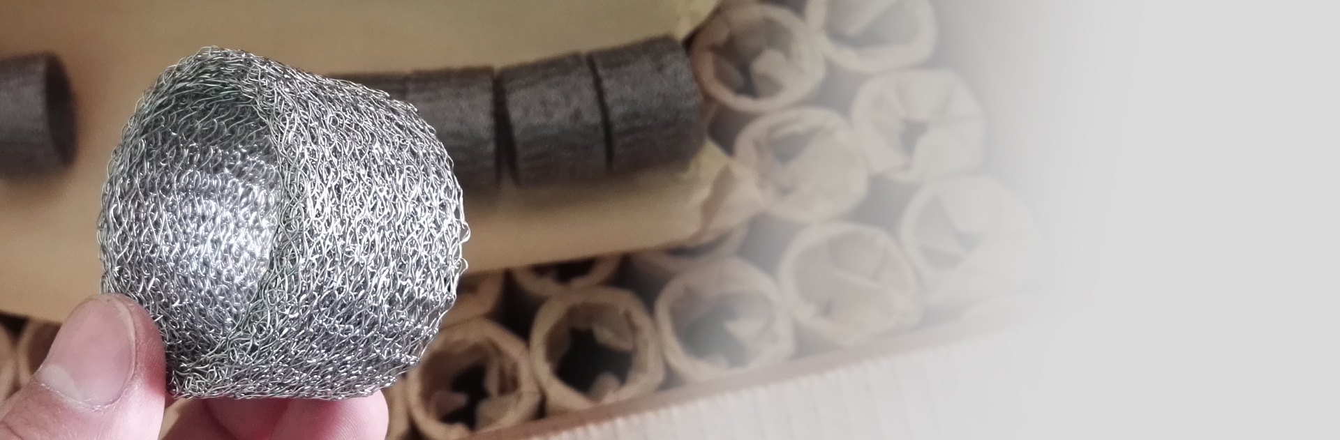 One hand is holding a compressed knitted wire mesh with ring shape, and other packs of meshes are in the carton box.